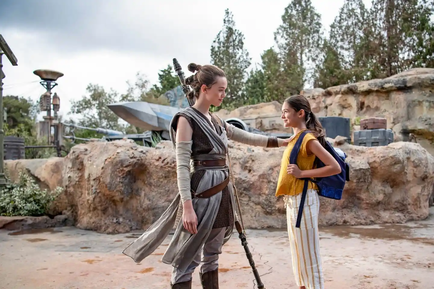 Rey meets a young girl in Star Wars: Galaxy&rsquo;s Edge with ships and spires in the background.