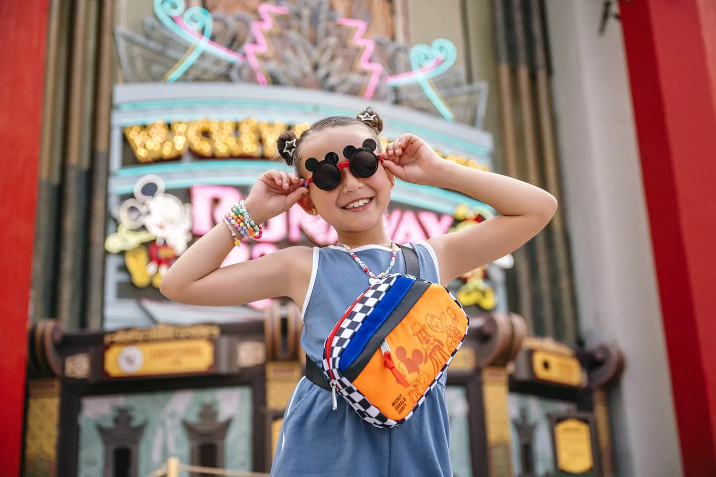 Young girl wearing Mickey sunglasses and Disney crossbody bag posing in front of the El CapiTOON Theater marquee for Mickey & Minnie&rsquo;s Runaway Railway.