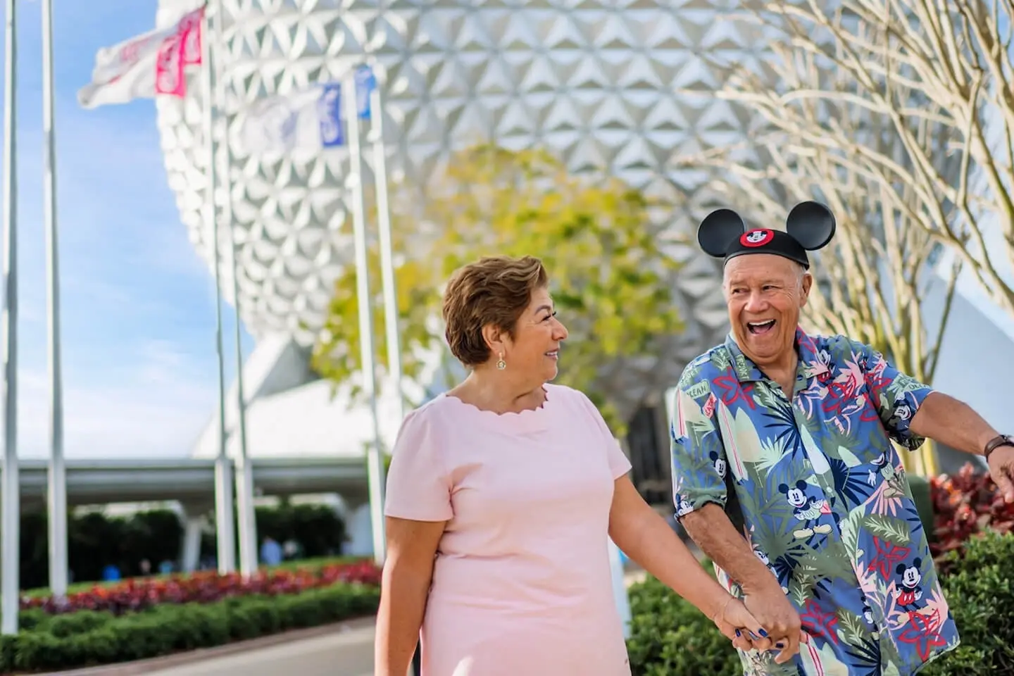 Couple holding hands in front of Spaceship Earth at EPCOT; man wearing Mickey ears and a Disney shirt.