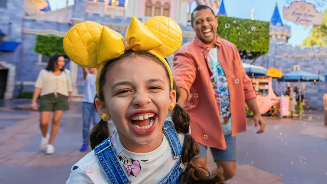A young girl wearing Minnie ears and smiling at the camera with her Dad behind her.